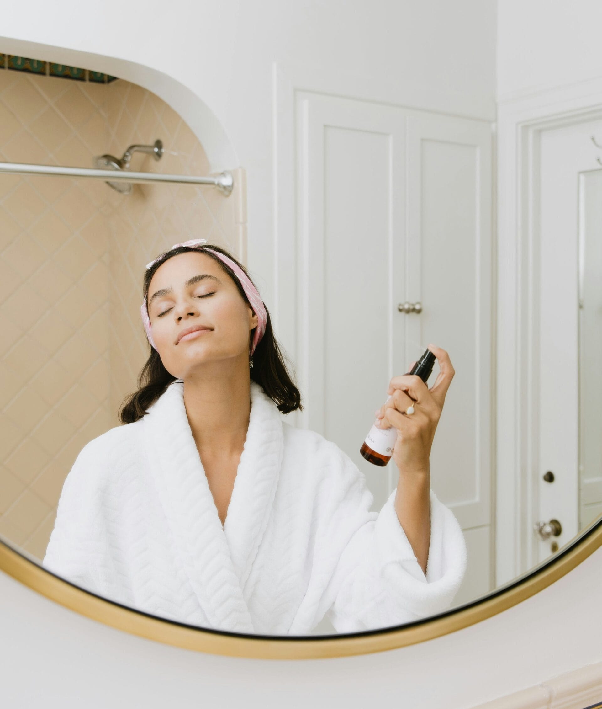 A woman in a robe in the bathroom spraying a mist on her face.